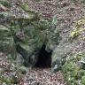 Das Foto zeigt eine von bemoosten Felsen eingerahmte kleine Höhle. Die Öffnung liegt am unteren Ende eines Waldhanges.