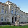 Blick auf die helle schlossähnliche Fassade des Naturkundemuseums in Karlsruhe mit Mittelteil und Seitenflügeln. Im vorkragenden Mittelteil befindet sich der Eingang zum Museum. Davor steht die Nachbildung eines Flugsauriers.