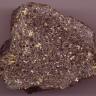 Nahaufnahme eines Gesteinsbrockens mit unregelmäßiger Form, violettgrau mit schwarzen, gelben und weißen Sprenkeln.