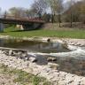 Blick auf einen schmalen Flusslauf mit flacher Staustufe und Strudelbildung im rechten Bildteil. Die Fließrichtung ist von links nach rechts. Der Fluß ist mit Steinufern befestigt. Im Hintergrund links ist eine Brücke, rechts ein Damm mit Bäumen sichtbar.