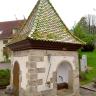 Blick auf ein gemauertes Brunnenhäuschen mit pyramidenförmigem Dach. Rechts ist eine Öffnung im Gebäude, dort ragt das steinerne Brunnenbecken heraus.