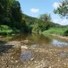 Blick auf ein teilweise trockenliegendes flaches Flussbett. Das Ufer rechts ist mit Gebüsch und Bäumen bewachsen. An der linken Uferseite steigen zusätzlich bewaldete Berghänge auf.