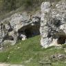 Seitlicher Blick auf eine an einem nach links geneigten Hang sich erhebende Höhle. Die Höhle besteht aus weißlichem, stark durchlöchertem Gestein. Eingänge finden sich rechts vorne sowie links hinten.