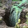 Das Bild zeigt einen von rechts oben kommenden Bachlauf, dessen grün eingefärbtes Wasser in eine sich im Vordergrund öffnende Erdspalte fällt.