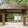 Blick auf ein großes, aus Holzpfosten gebautes und überdachtes Tor, das zur Aachquelle führt. Im Hintergrund sind ein Teil des Quellsees, das angrenzende belaubte Ufer sowie Hinweistafeln zu erkennen.