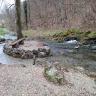 Von einer Ortschaft links hinten strömt ein Wasserlauf nach rechts am Betrachter vorbei. Links der Bildmitte umfließt er dabei eine kleine, von Felsgestein umrandete Insel mit Baum und Sitzbank. Das vordere Ufer ist flach, das hintere bildet ein Waldhang.