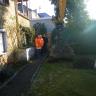 Das Foto zeigt ein blassgelbes Haus mit Garten. Ein Bagger hebt ein Loch aus. Mehrere Personen in orangefarbenen und dunklen Jacken stehen am Rand und schauen dem Bagger zu.
