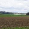 Über dunkelbraune Äcker im Vorder- und Mittelgrund geht der Blick über flachwelliges Gelände mit grünen Hügeln und Wiesen. Im Hintergrund ziehen sich langgestreckte bewaldete Bergrücken hin.
