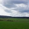 Blick über eine flachhügelige Wiesenlandschaft, die im Hintergrund von Ackerstreifen unterbrochen sowie von bewaldeten Bergrücken begrenzt wird. Entlang der Bergrücken rechts sind mehrere Windräder zu erkennen. Im Vordergrund verläuft eine schmale Straße.