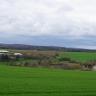 Blick über einen zum Mittelgrund hin abfallenden Wiesenhang. Dahinter, in einer Senke, liegt ein verbuschter Streifen mit Bäumen und Gebäuden. Weiter zum Hintergrund hin folgen flache Wiesen und Äcker sowie ein langer bewaldeter Bergrücken.