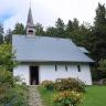 Blick auf eine kleine Kapelle mit weißen Wänden und hellblauem Dach. Ein aus Steinplatten gelegter Weg, gesäumt von blühenden Hecken, führt hinauf zum Gebäude. Im Hintergrund steht dichter Wald.