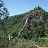 Blick aus großer Höhe auf einen zum Hintergrund hin aufsteigenden, bewaldeten Bergkegel. Der obere Teil des Kegels zeigt Felsformationen, zudem verläuft auf der linken Seite, in der Bildmitte, ein vorspringender, mauerartiger Felsengang.