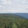 Blick von erhöhtem Standort über weite bewaldete Bergrücken, die rechts in ein Tal münden. Im fernen Hintergrund dehnen sich weitere bewaldete Höhenzüge aus.