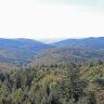 Blick aus großer Höhe über Baumwipfel auf mehrere Reihen bewaldeter Berge. Zwischen den Bergrücken sind links und rechts Taleinschnitte erkennbar. Im fernen Hintergrund, über der Bildmitte, öffnet sich eine weite Ebene.