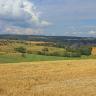 Blick über eine hochgelegene, abgeerntete Ackerfläche. Dahinter, tiefer, liegen weitere Äcker, Wiesen und Waldstücke. Im Hintergrund sind bewaldete Höhenzüge erkennbar.