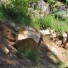 Das Bild zeigt einen nach links ansteigenden Hang, der im unteren Teil von größeren Felsbrocken durchsetzt ist. Braunes Bodenmaterial hat die Felsen teilweise überdeckt. Oberhalb der Felsen ist Pflanzenbewuchs und dahinter weiteres Felsgestein erkennbar.