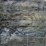 Teilansicht einer etwa 15 m hohen Gesteinswand. Das Gestein ist dunkelgrau bis braun und weist eine schrundige Oberfläche sowie eine feine Schichtung auf. Rechts unten lehnt eine Messlatte.