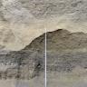 Nahaufnahme der Abbauwand einer Kies- und Sandgrube. In der Mitte der Wand verläuft eine gebogene Linie, die hellbraune Schichten oben von dunkelgrauen unten trennt. Eine Messlatte reicht bis zum höchsten Punkt des Bogens.
