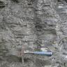 Blick auf eine graue Gesteinswand mit waagrecht verlaufenden feinen Furchen und Scherben. Unten mittig zeigt ein Hammer die Größenverhältnisse an.