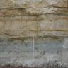 Blick auf eine Steinbruchwand mit verschiedenen Lagen. Die unterste Lage ist grau mit bläulichen Streifen. Die mittlere und oberste Lage ist jeweils hellbraun bis grau mit orangen Streifen und rötlichen Flecken. Die dritte Lage ist zudem dünner gebankt.