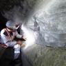 Ein Bergbauarbeiter links im Bild beschäftigt sich mit einer Wand aus großen, weißlichen Kristallen, die auf grünlichem Gestein aufliegt.