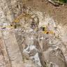 Blick in einen Steinbruch: Das hellbeige bis hellgraue Gestein ist stark geklüftet, dabei stehen die Klüfte sehr steil. Zwischen den Felswänden befindet sich - halb verdeckt - ein gelber Bagger.