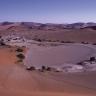 Das Bild zeigt eine afrikanische Wüste mit Dünen im Hintergrund und einem gewundenen, beinahe ausgetrockneten Fluss im Vordergrund. Der Fluss endet in einem runden, flachen Becken.