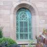 Blick auf ein Rundbogenfenster mit blaugrünen Sprossen und Verzierungen. Das Fenster ist von rötlich grauen Mauersteinen umgeben.