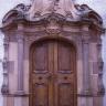 Das Foto zeigt das Eingangsportal einer Kirche mit hölzernen Türen und umlaufenden Verzierungen aus rötlich grauem Stein.