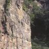 Blick auf eine hoch aufragende, aus rötlich grauem Gestein bestehende Abbauwand eines Steinbruches. Ein Mensch rechts unten begutachtet die Steinwand, die nach oben hin von Pflanzen bewachsen ist.