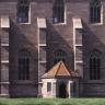 Das Bild zeigt die rötlich graue Fassade einer Kirche oder eines Klosters mit zwei Fensterreihen und vier senkrechten Stützpfeilern. Etwas davor steht ein niedriges Türmchen mit rotem Spitzdach.