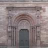 Eingangsportal einer Kirche mit Doppeltüre, schmalen Säulen und Verzierungen. Das verwendete Gestein ist von rötlich grauer Farbe.