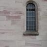 Das Foto zeigt ein schmales vergittertes und oben abgerundetes Fenster in einer rötlich grauen Steinmauer.