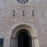 Frontansicht des Eingangs einer Kirche. Das dunkle Eingangstor wird von schmalen Säulen und runden Bögen umrahmt. Die Kirche ist aus rötlich grauen Steinen errichtet. Oben ist noch eine Fenster-Rosette zu sehen.
