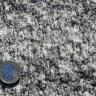 Nahaufnahme von weißem Gestein mit kristalliner Oberfläche und dunkler Marmorierung. Links unten dient eine Euro-Münze als Größenvergleich.
