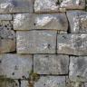 Das Bild zeigt eine Trockenmauer aus rechteckigen, hellgrauen Gesteinsblöcken. Die Blöcke haben alle eine leicht unterschiedliche Form. Manche Mauerfugen sind bewachsen.