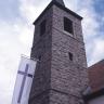 Blick von unten auf einen viereckigen Kirchturm mit spitzem Dach. Der Turm ist aus rötlichem Gestein gebaut. Auf der linken Seite des Turms ist eine weiße Fahne mit rotem Kreuz angebracht.