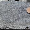 Nahaufnahme eines grauen Steinbrockens mit unebener Oberfläche und kleineren schwarzen Sprenkeln. Rechts oben dient eine Cent-Münze als Größenvergleich.