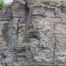 Blick auf eine weißlich graue, von Rissen und Nischen durchzogene Steinbruchwand. Der Sockel der Wand besteht aus gröberen Blöcken; nach oben hin sind die Lagen plattig. Unter der bewachsenen Kuppe sind Rottöne erkennbar.