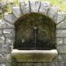 Blick auf einen steinernen Brunnen mit rechteckigem Becken und steinerner Rückwand. Der Brunnen wird auf allen Seiten von grauen Mauersteinen eingefasst, die oben im Bogen angeordnet sind.