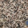 Nahaufnahme einer hellen Gesteinsoberfläche mit schwarzen und rötlichen Sprenkeln. Rechts unten dient eine Euro-Münze als Größenvergleich.