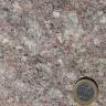 Nahaufnahme einer grobkörnigen, kristallinen Gesteinsoberfläche, Farbe rötlich grau mit größeren hellen Einschlüssen. Rechts unten liegt eine Euro-Münze.
