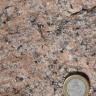 Nahaufnahme einer kristallinen Gesteinsoberfläche, rötlich braun bis grau mit dunkleren Sprenkeln. Rechts unten dient eine Euro-Münze als Größenvergleich.