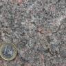 Nahaufnahme einer kristallinen Gesteinsoberfläche, dunkelgrau mit rötlichen und vereinzelt weißen Einschlüssen. Links unten liegt eine Euro-Münze.