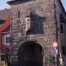Das Foto zeigt ein altes gemauertes Stadttor als Turmbau mit Dach und bogenförmigem Durchlass sowie angebautem Erker. Die Mauersteine auf der Frontseite sind rötlich grau.