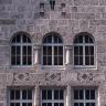 Ansicht einer hohen Hausfassade aus rötlich grauem Mauerwerk mit zwei Fensterreihen, einem Giebelfenster und kleineren Verzierungen.