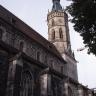 Teilansicht einer Kirche mit Haupt- und Seitenschiff aus dunkelgrauem Gestein sowie Turm (rechts im Bild) aus gelblich grauen Steinen.