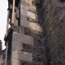 Teilansicht einer Kirchenwand mit linksseitig angebautem Strebepfeiler aus dunkelgrauen Mauersteinen. Teilweise sind auch hellgraue Steine verbaut.