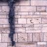 Nahaufnahme einer Wandverkleidung mit rötlich grauen Steinplatten, ähnlich Kacheln, in unregelmäßigem Muster. Links hängt ein stilisiertes, dunkelgraues Metallkreuz an der Wand.