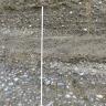 Teilansicht einer Gesteinswand mit (von oben): feinem Kies, eingebettet in Sand, einer glattgestrichenen Schicht sowie gröberem Kies, der ebenfalls von Sand umlagert ist. Links ist ein Zollstock angelehnt.